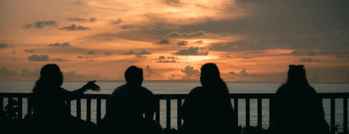 Four people sitting together in silhouette, talking as the sun sets over the sea