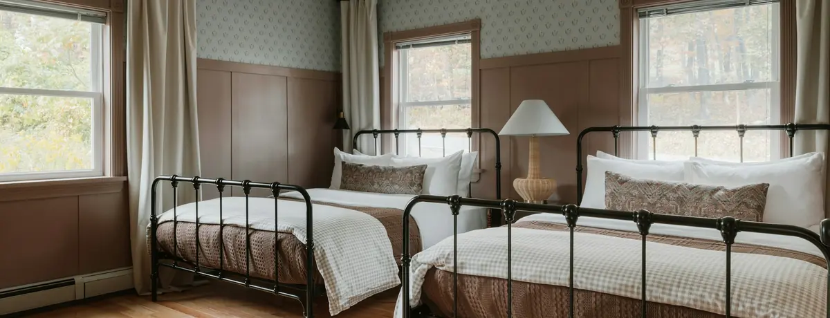 A simple guest room with two made-up beds and daylight coming through three windows